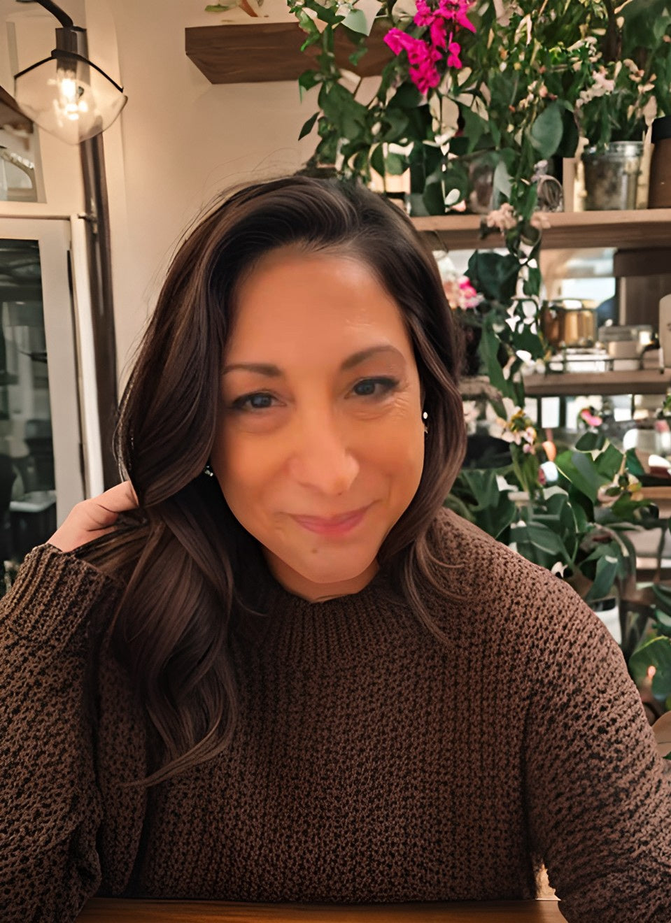 Image of Juliette Aiyana in brown sweater. Behind her are shelves with plants and a lamp.